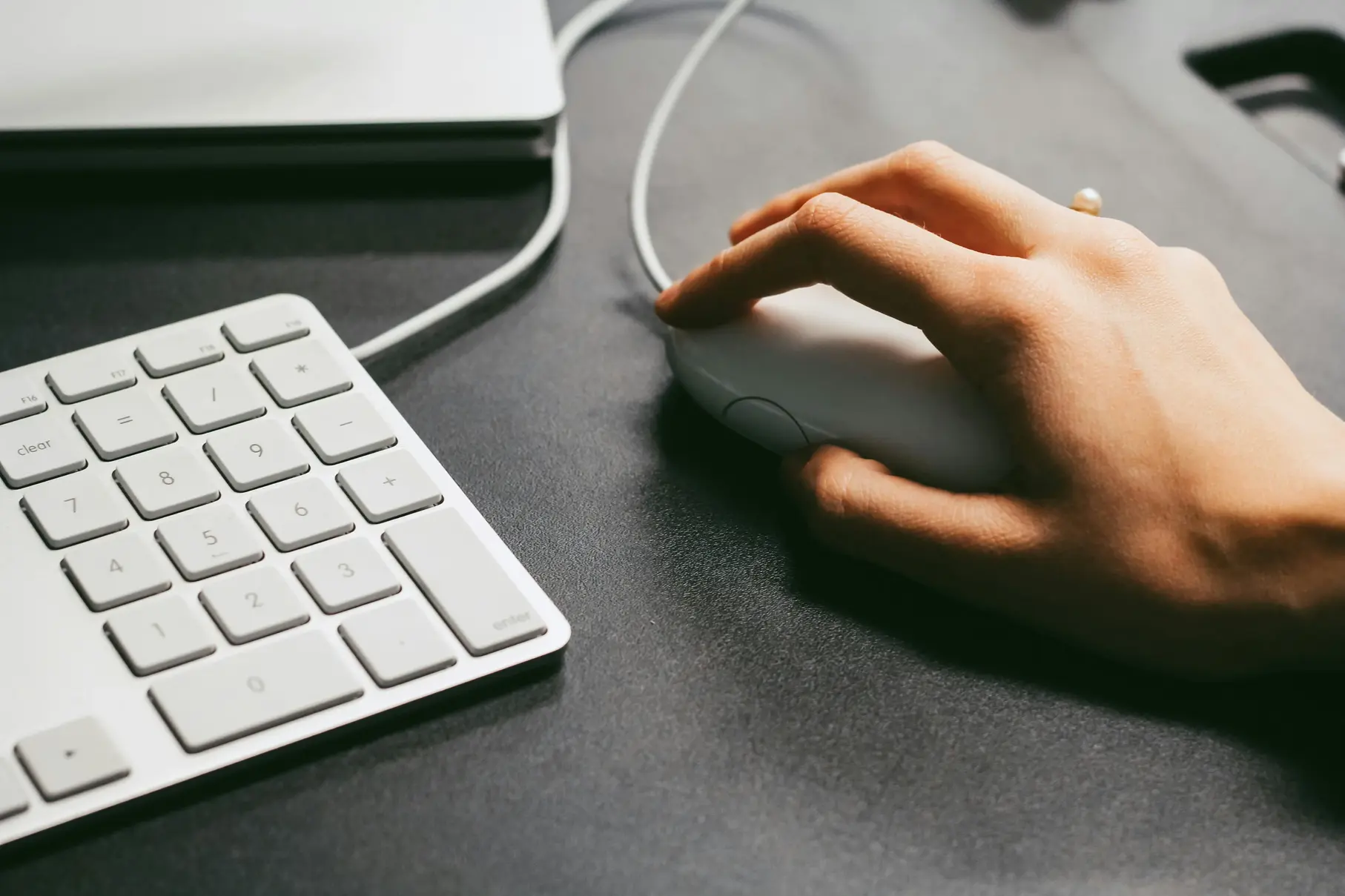 Close-up of hand using computer mouse next to white keyboard on dark office desk.