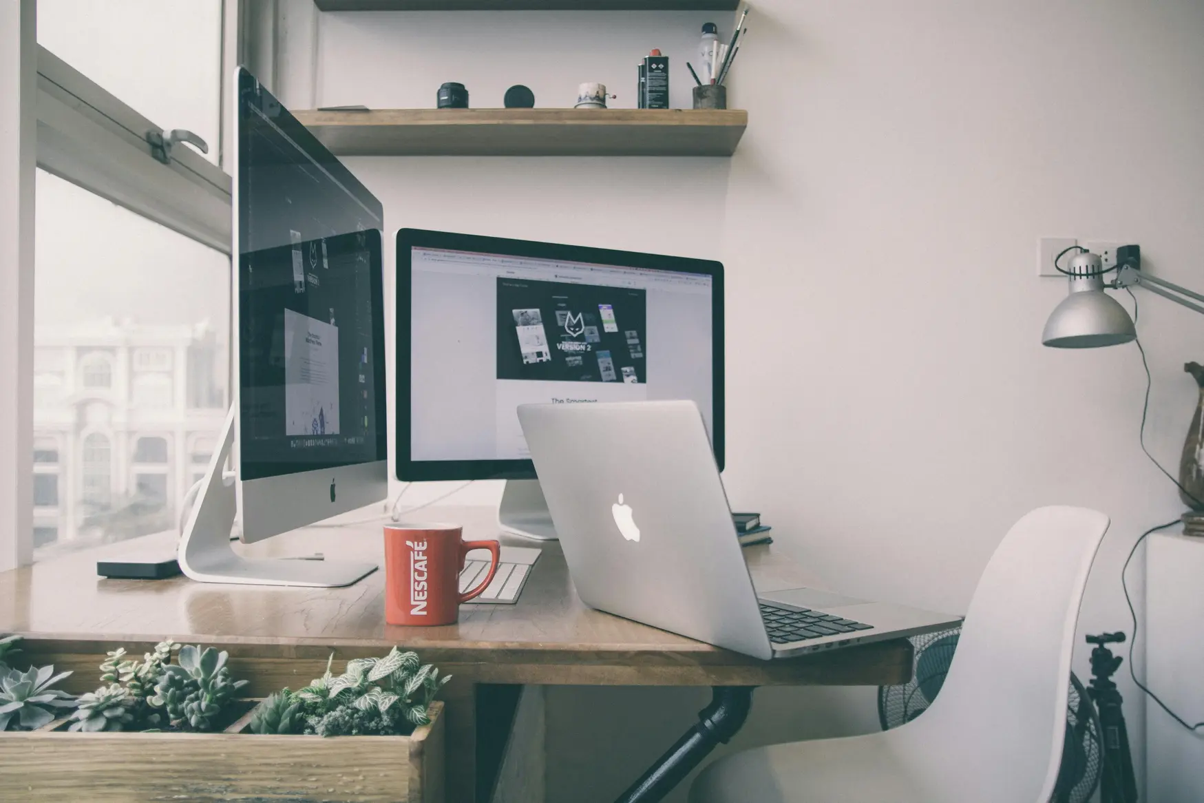 Modern home office setup with iMac, MacBook, red Nescafé mug, and indoor plants on a wooden desk