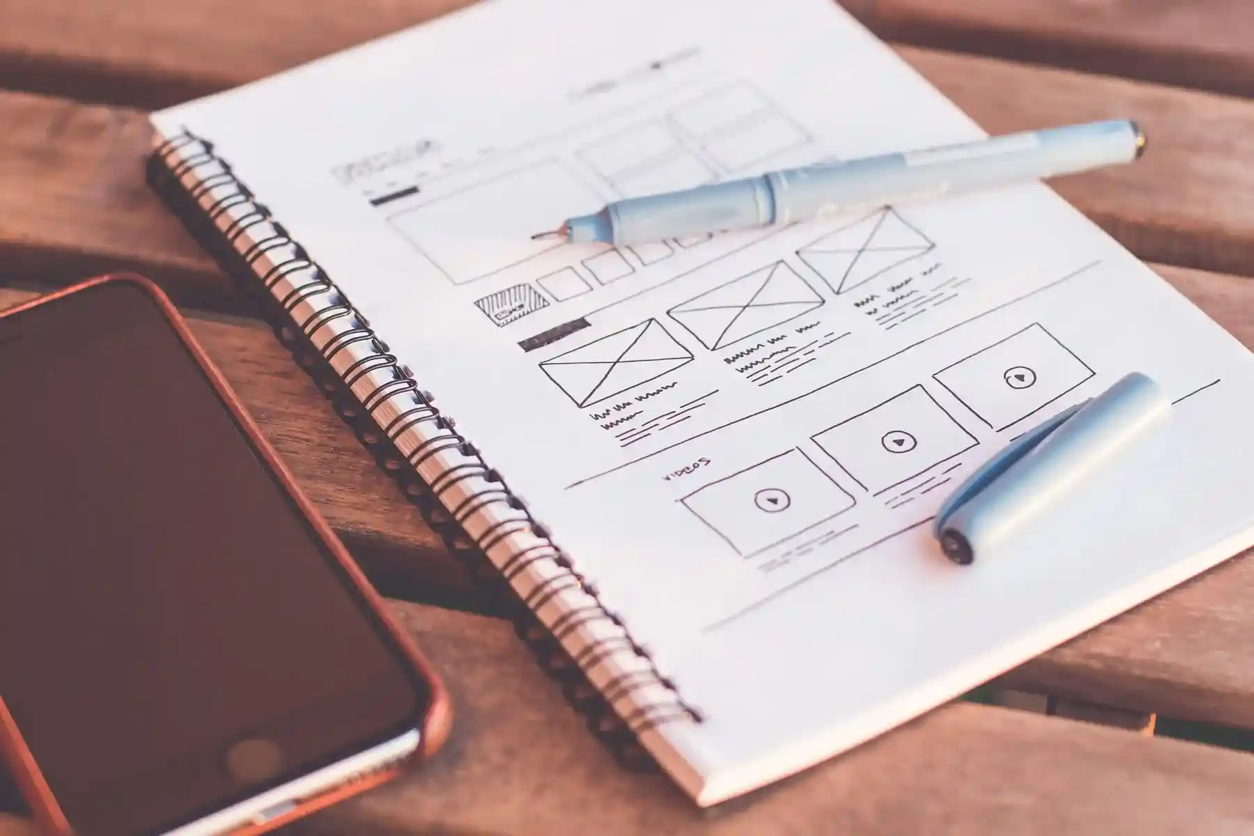 Notebook with website wireframe sketches, blue pen, and smartphone on a wooden desk